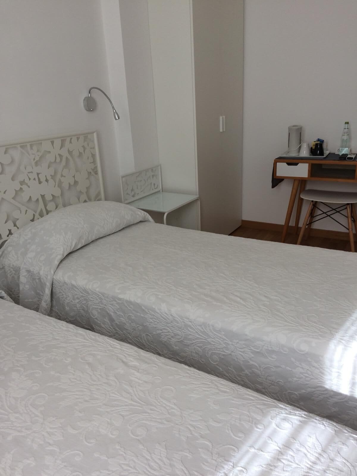 A hotel room with two beds, a desk, a chair, and a wall-mounted lamp.
