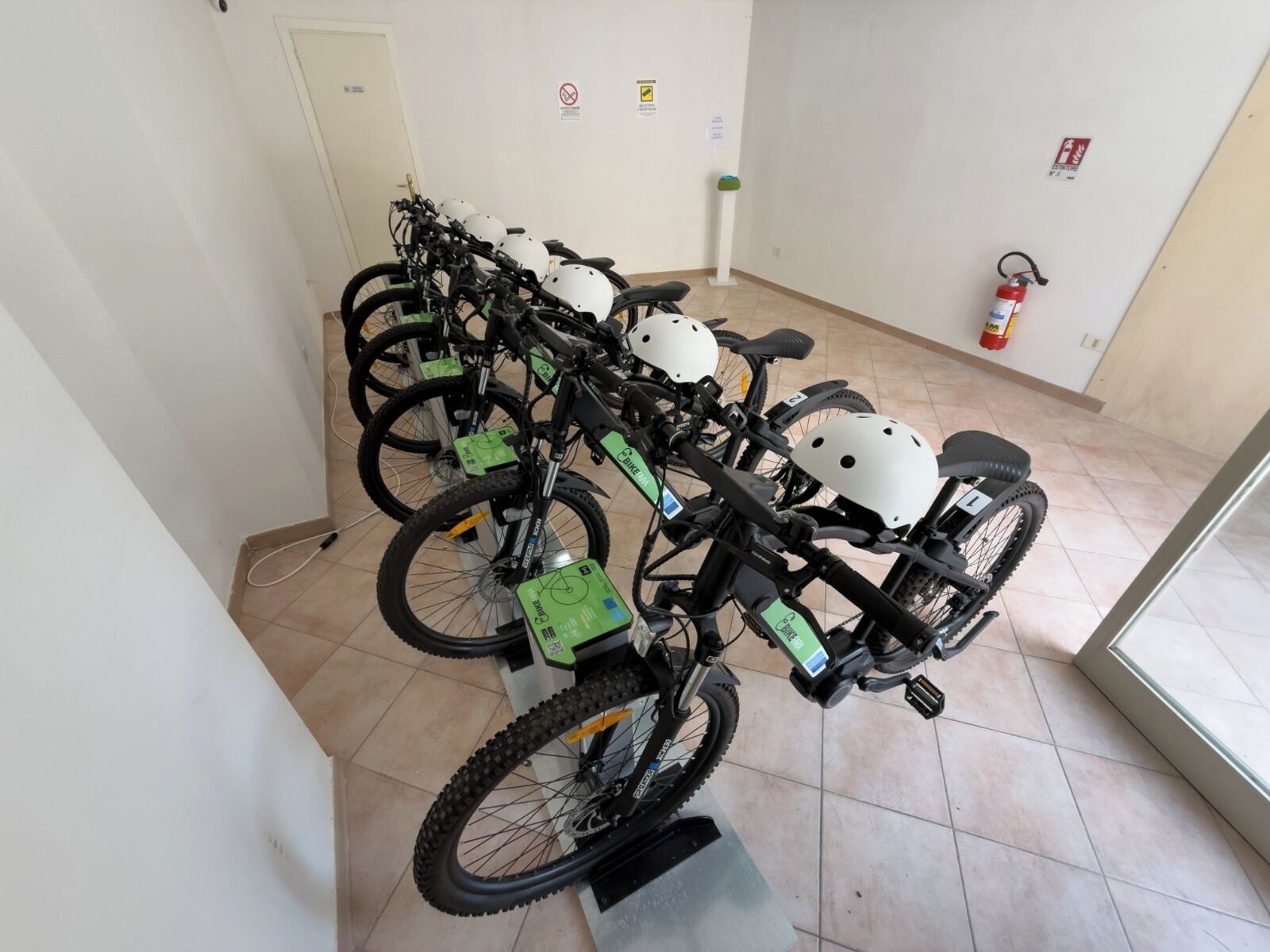 Une rangée de vélos, chacun équipé d’un casque et d’une étiquette verte, une porte blanche et un extincteur accroché au mur.
