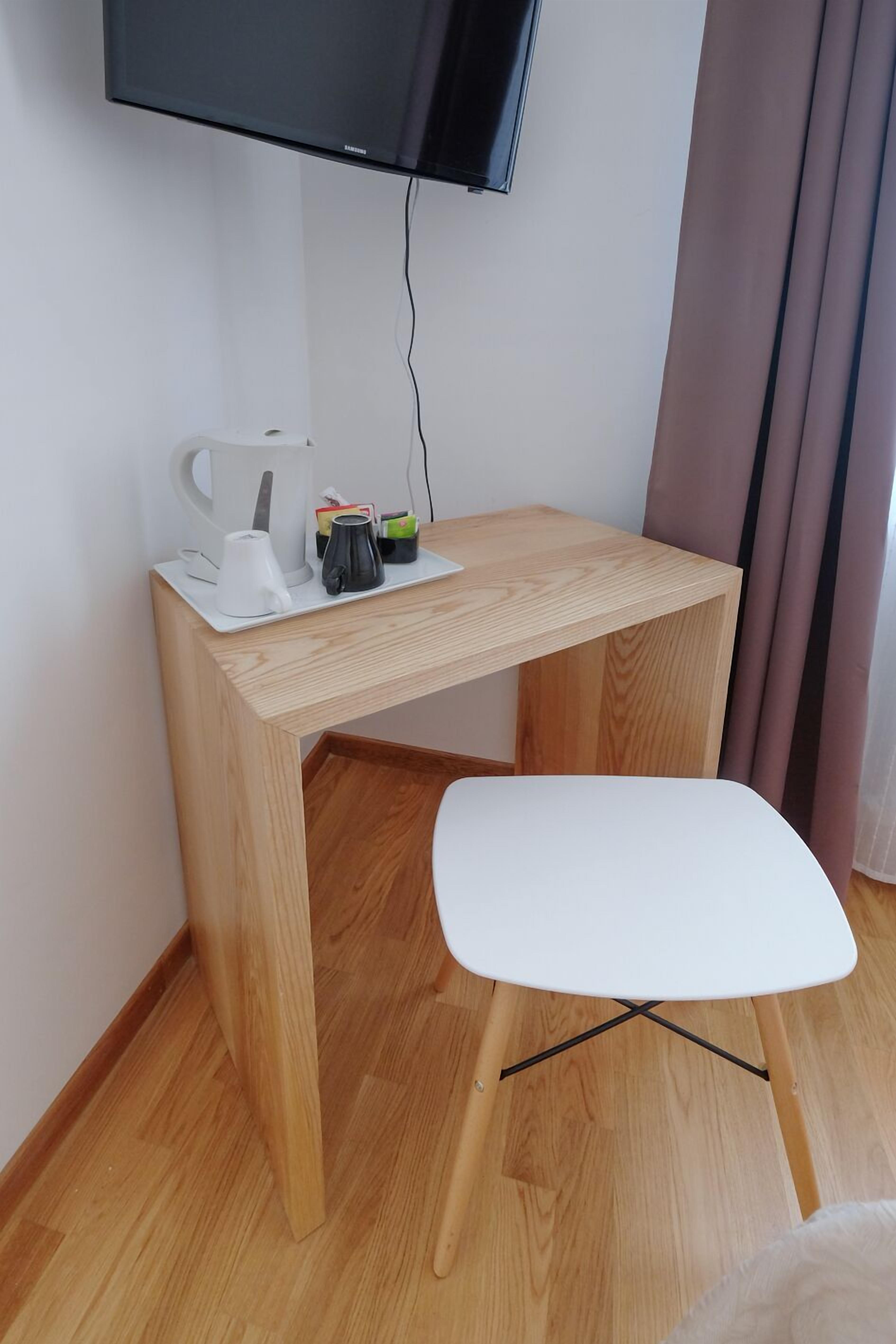 Una habitación pequeña y moderna con un escritorio de madera, una silla blanca, un televisor montado en la pared y una cafetera sobre el escritorio.
