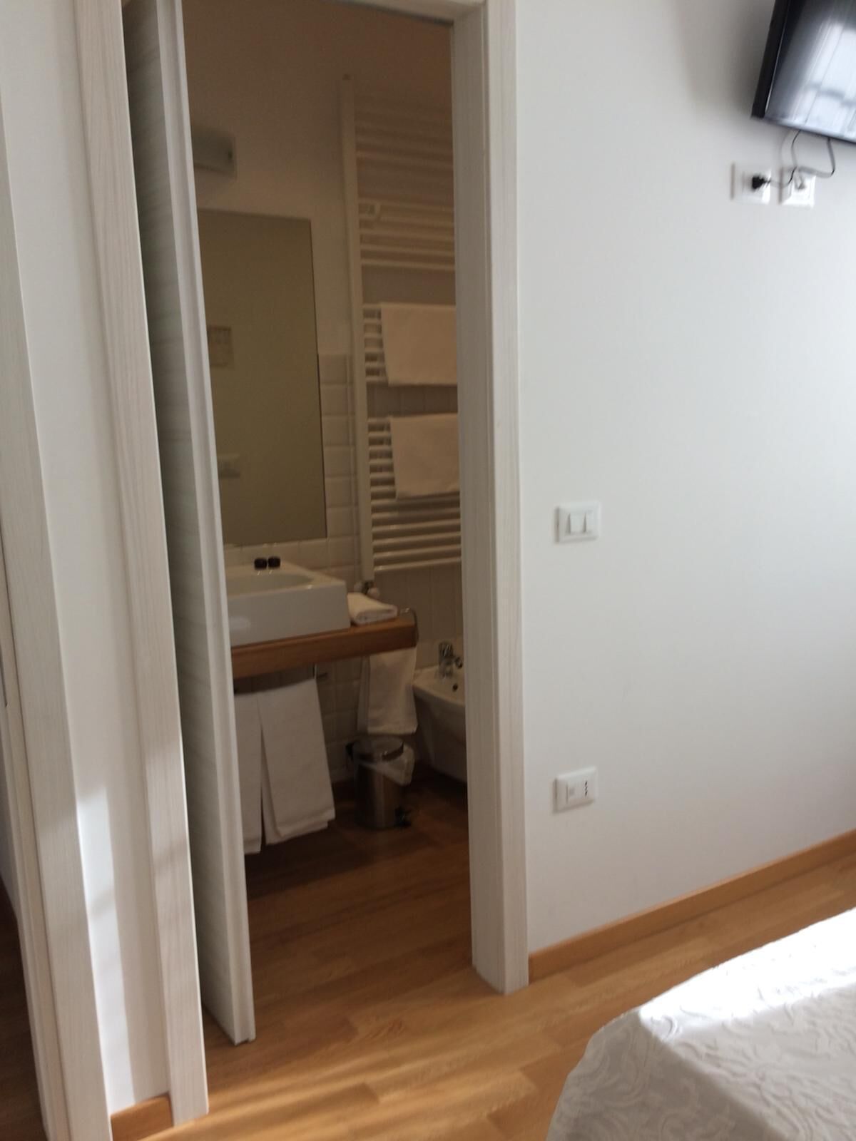 Een slaapkamer met een houten vloer, een bed, een aan de muur bevestigde televisie en een badkamer zichtbaar door een geopende deur.