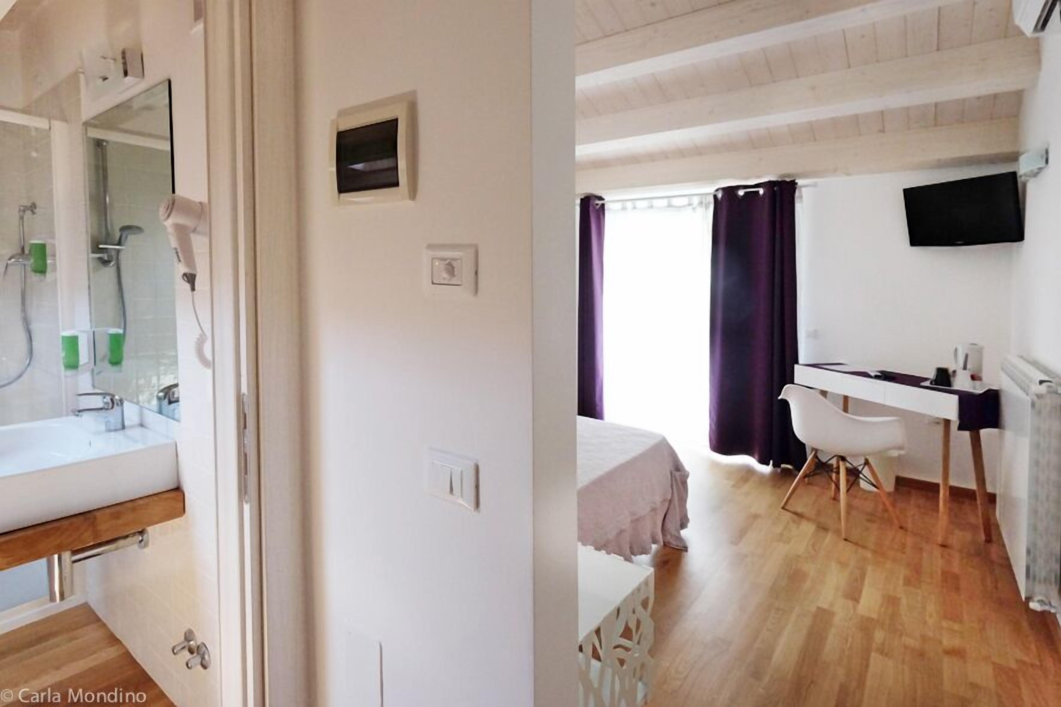 A room with a wooden floor, a bed with a white headboard, a desk with a chair, a TV mounted on the wall, and a window with purple curtains.