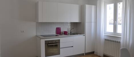 Cocina compacta con armarios blancos, horno de acero inoxidable, lavamanos y ventana con cortinas.