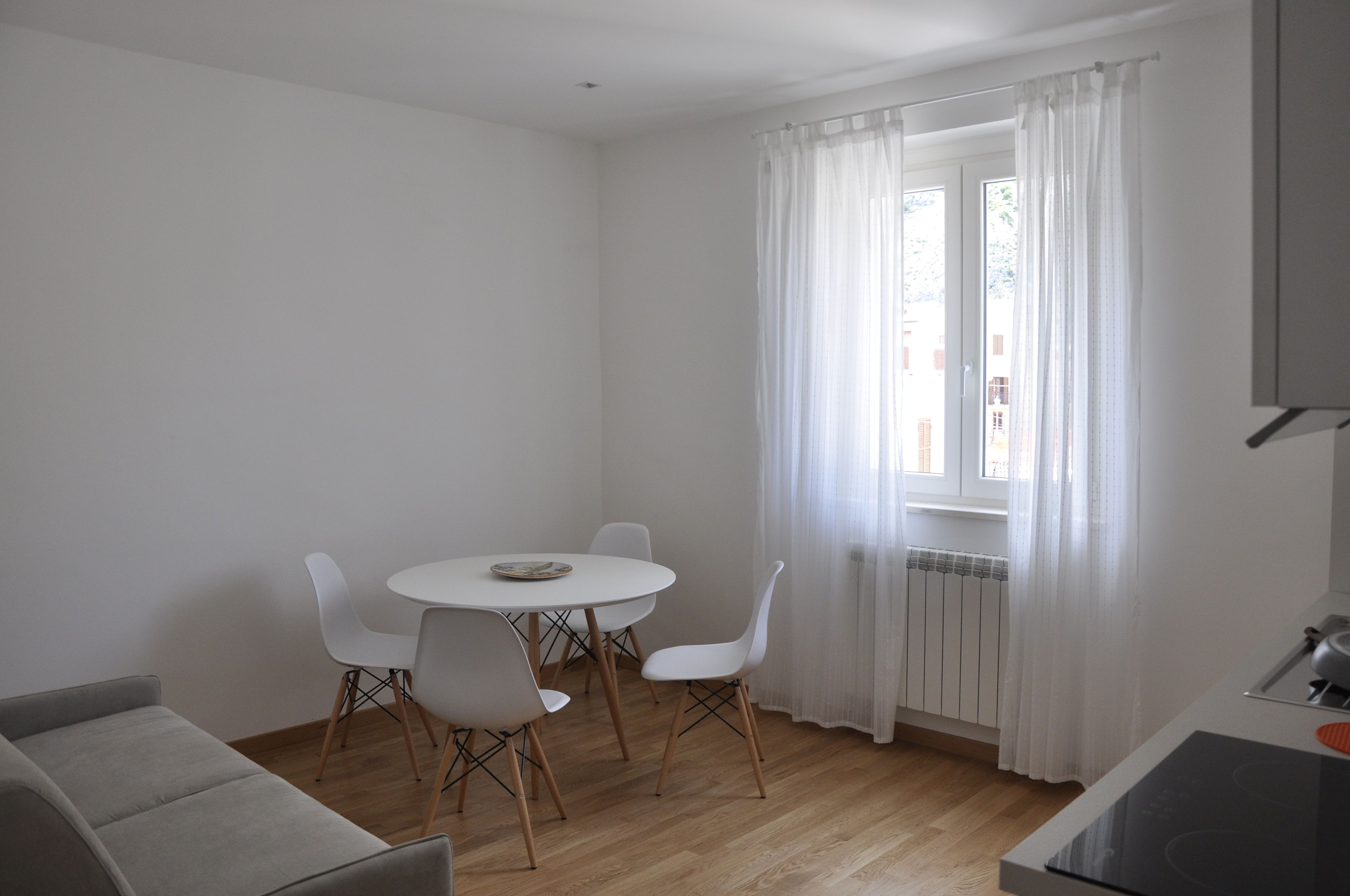 Una habitación moderna y minimalista con una mesa de comedor redonda, cuatro sillas blancas, un sofá gris y una ventana con cortinas de gasa.