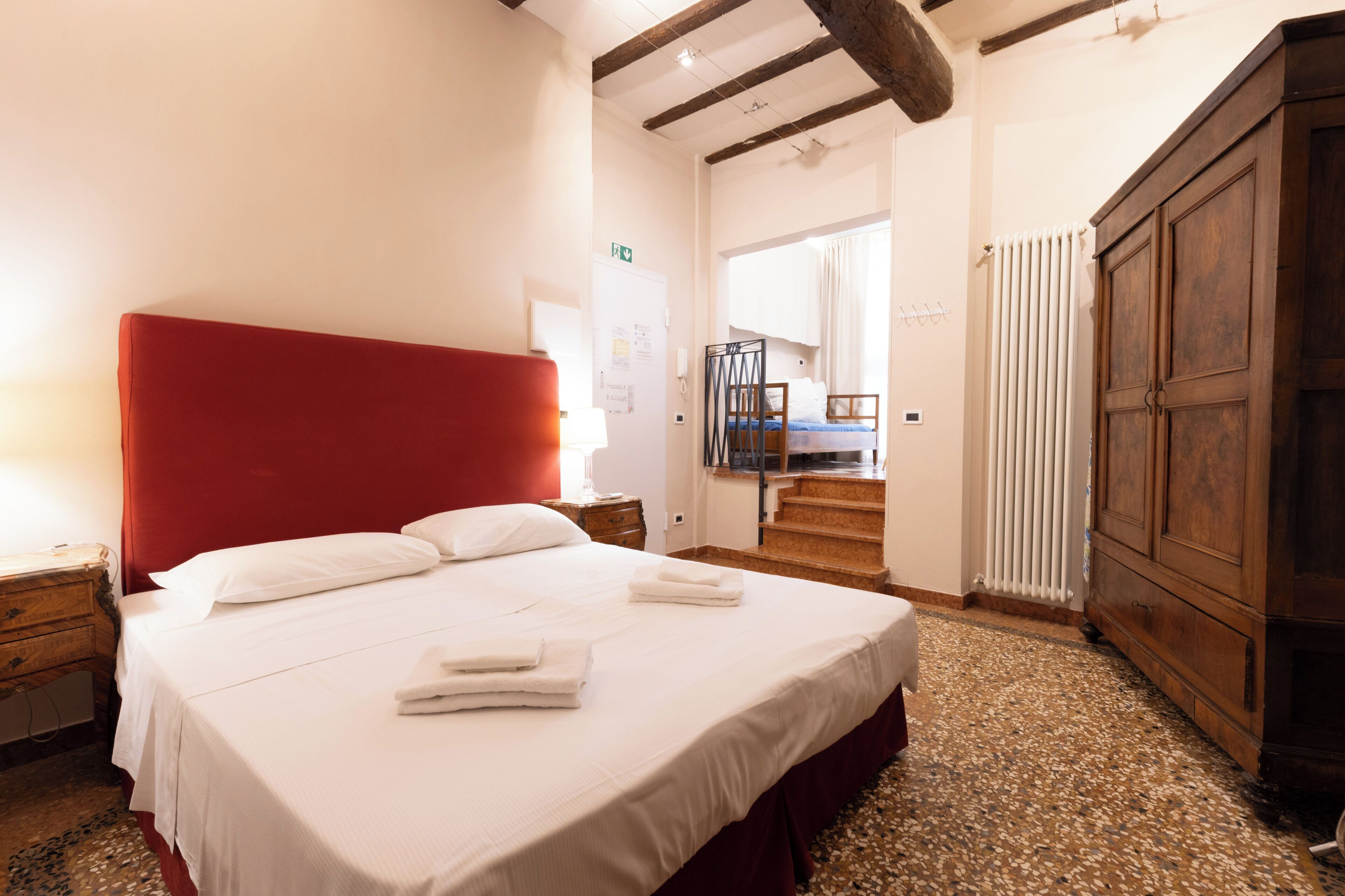 A hotel room with a large bed, a wooden wardrobe, a radiator, and a staircase leading to another room.