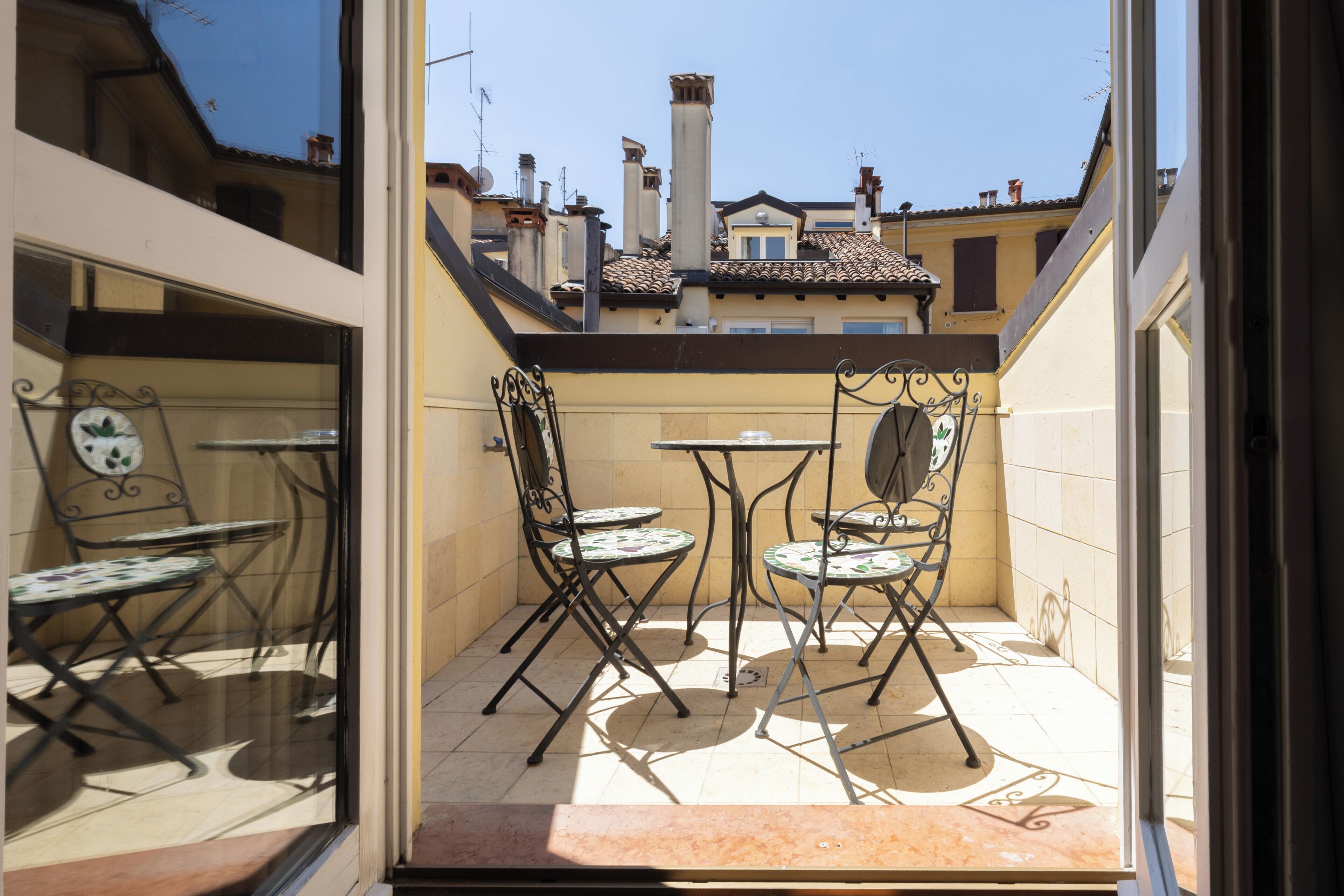 Eine kleine Terrasse mit zwei Tischen und vier Stühlen, ein Fenster mit einem dekorativen Spiegel und Blick auf benachbarte Gebäude.