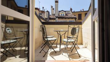 A small patio with two tables and four chairs, a window with a decorative mirror, and a view of neighboring buildings.