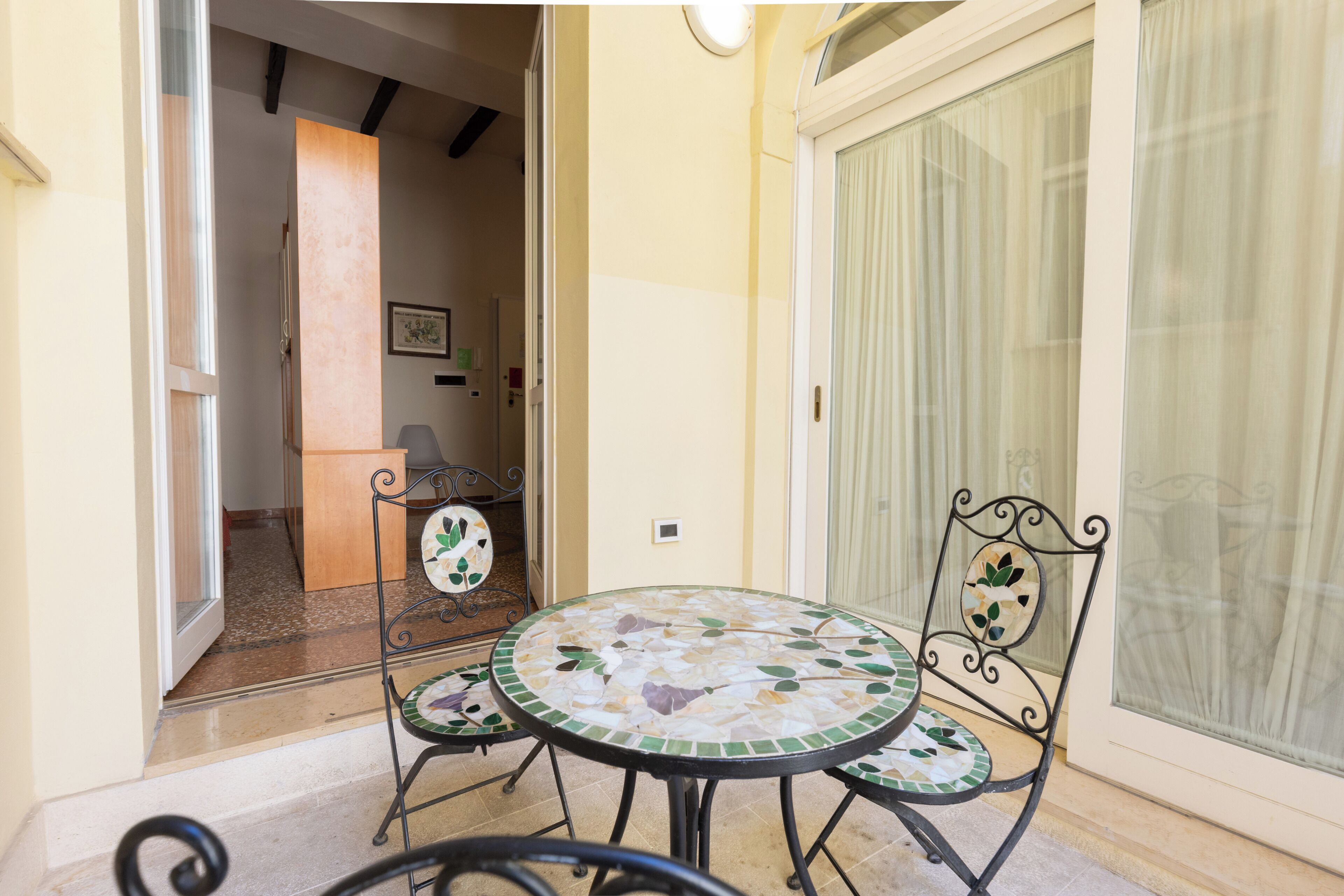 A small patio area with a round table and two chairs, a door, and a window with curtains.