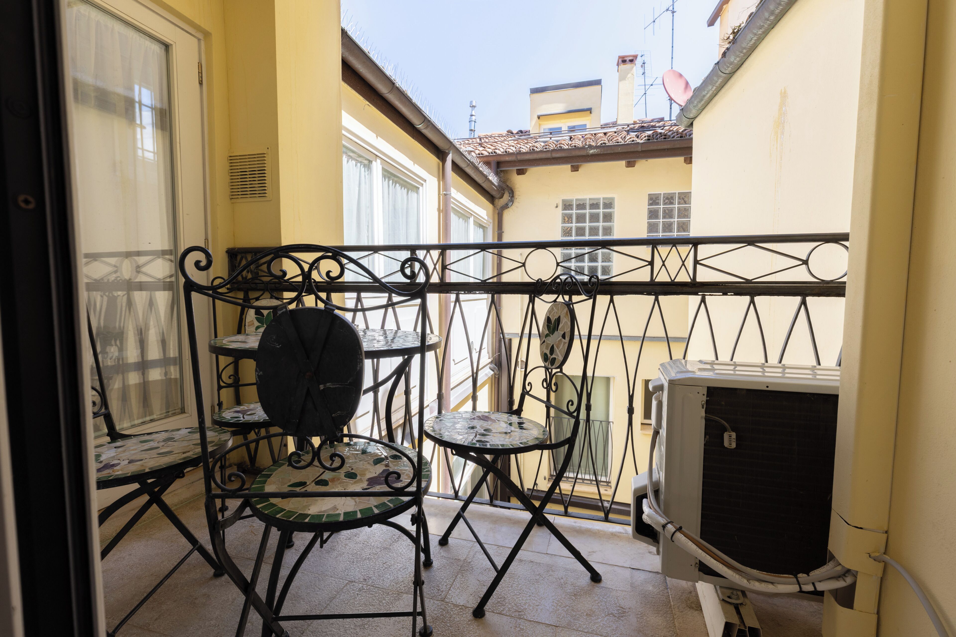 A balcony with a small table and chairs, an air conditioning unit, and a view of the street.