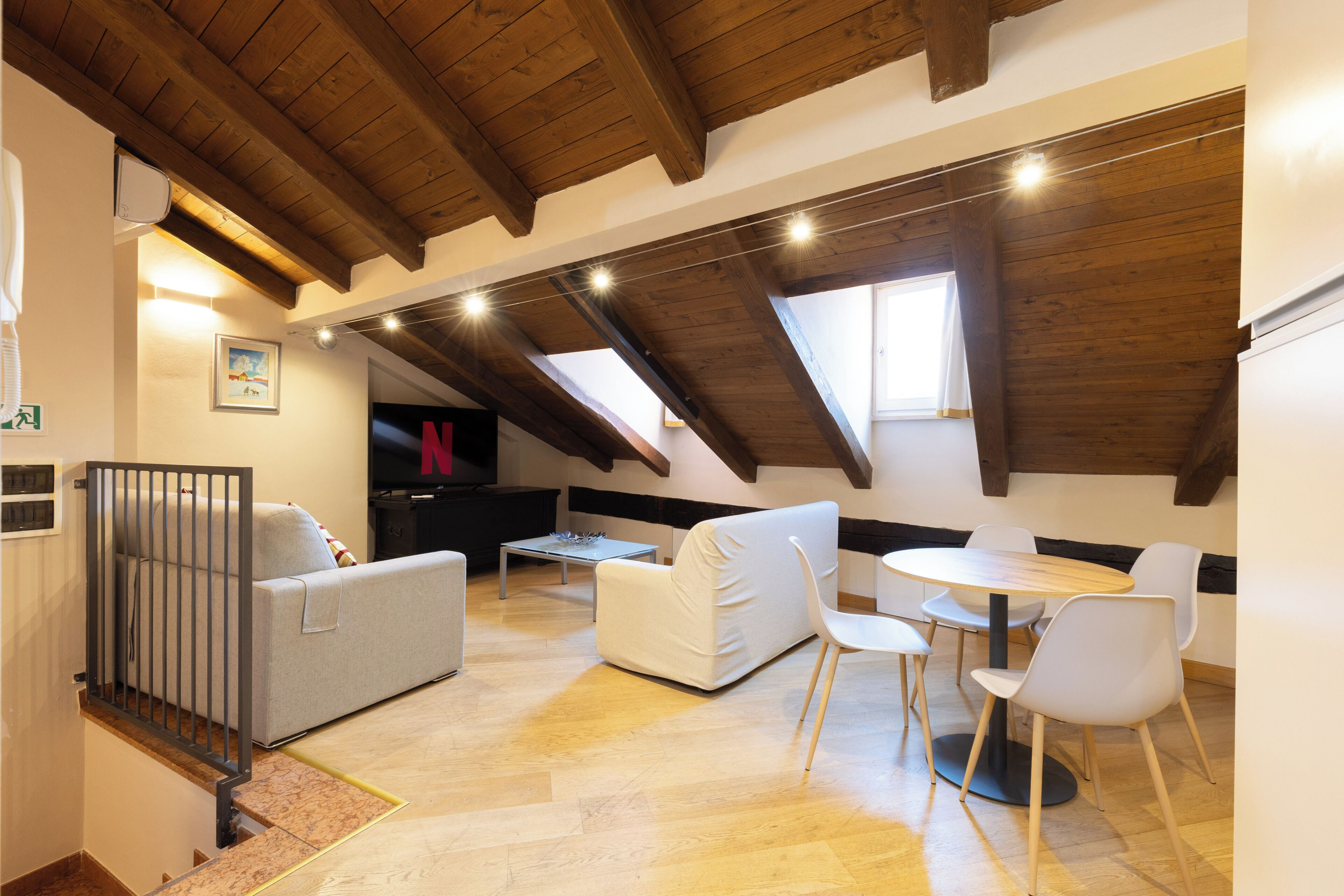 A loft-style room with a high ceiling, wooden beams, a sofa, a dining table with chairs, and a television.