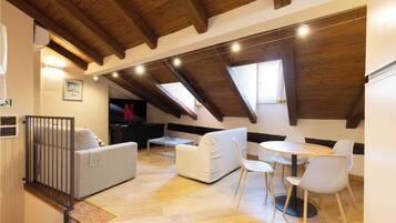 A loft-style room with a high ceiling, wooden beams, a sofa, a dining table with chairs, and a television.
