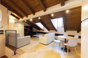 A loft-style room with a high ceiling, wooden beams, a sofa, a dining table with chairs, and a television.