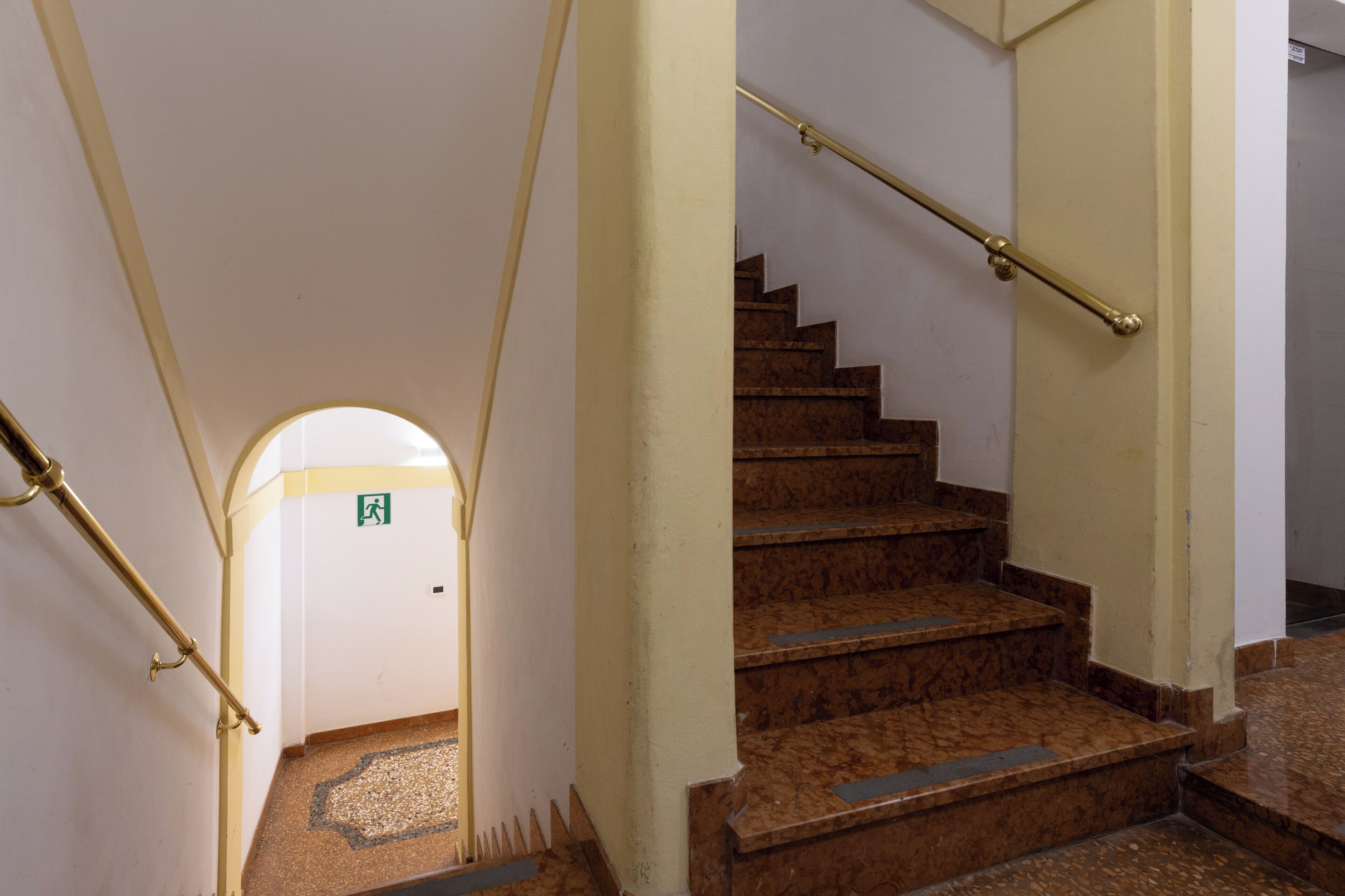 A staircase with a handrail, a patterned floor, and a door with an emergency exit sign.