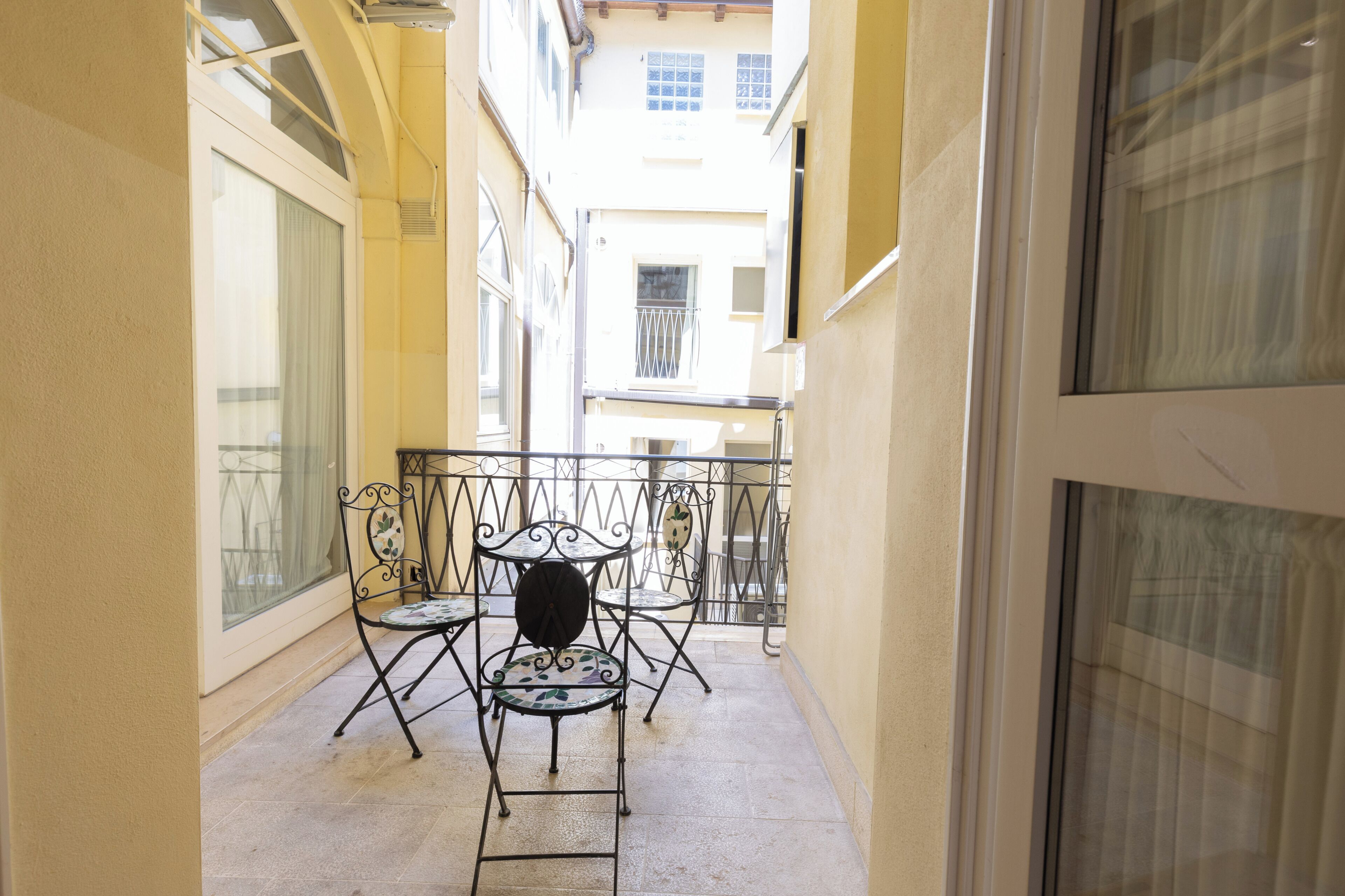 A narrow balcony with metal chairs and a table, overlooking a street.