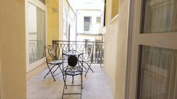 A narrow balcony with metal chairs and a table, overlooking a street.