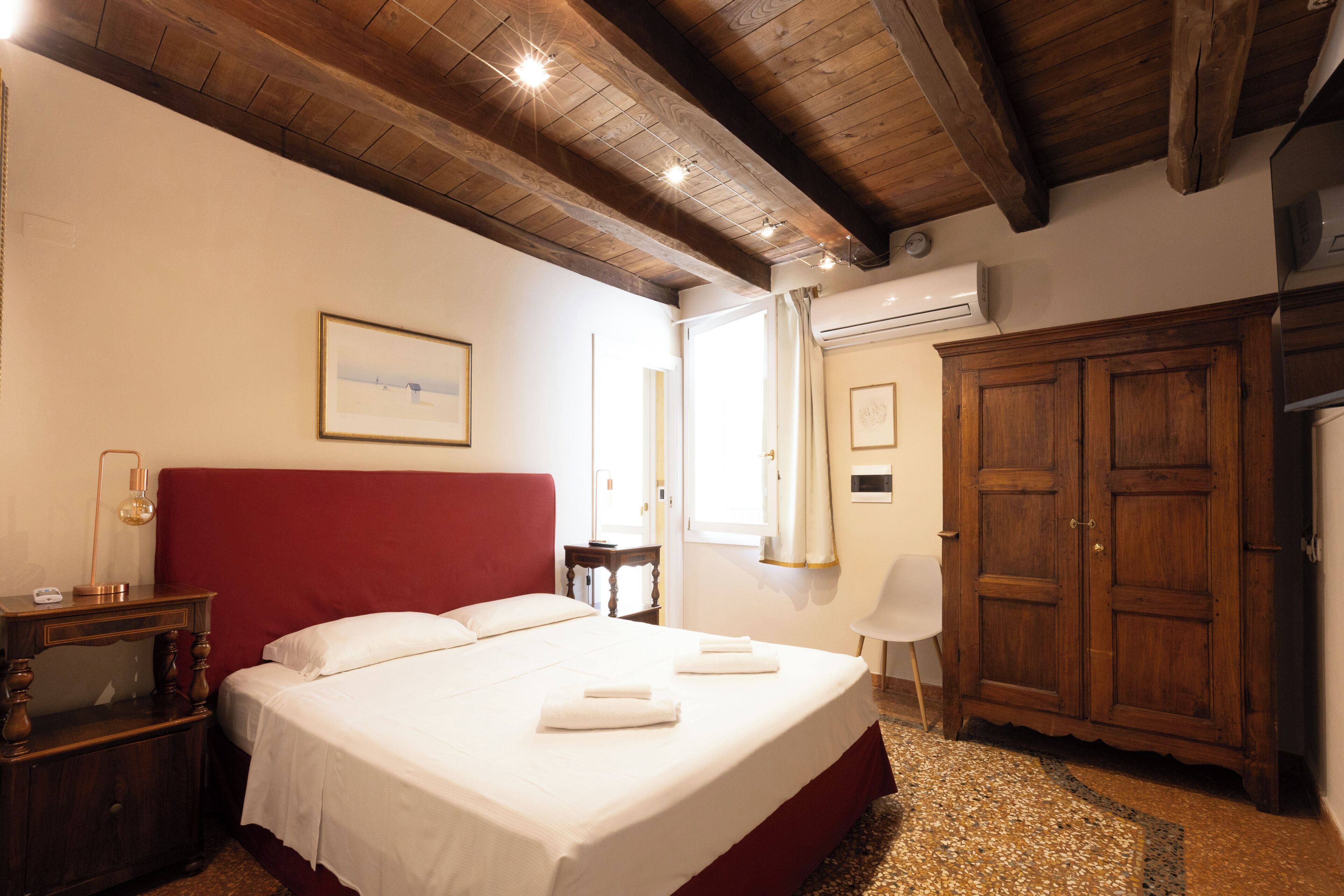 A hotel room with a large bed, a wooden wardrobe, a desk with a lamp, and a window with curtains.