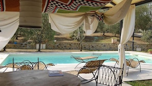 Ein Poolbereich mit Holztisch und Metallstühlen, einem karierten Sonnensegel und Blick auf einen Swimmingpool und Grünflächen.