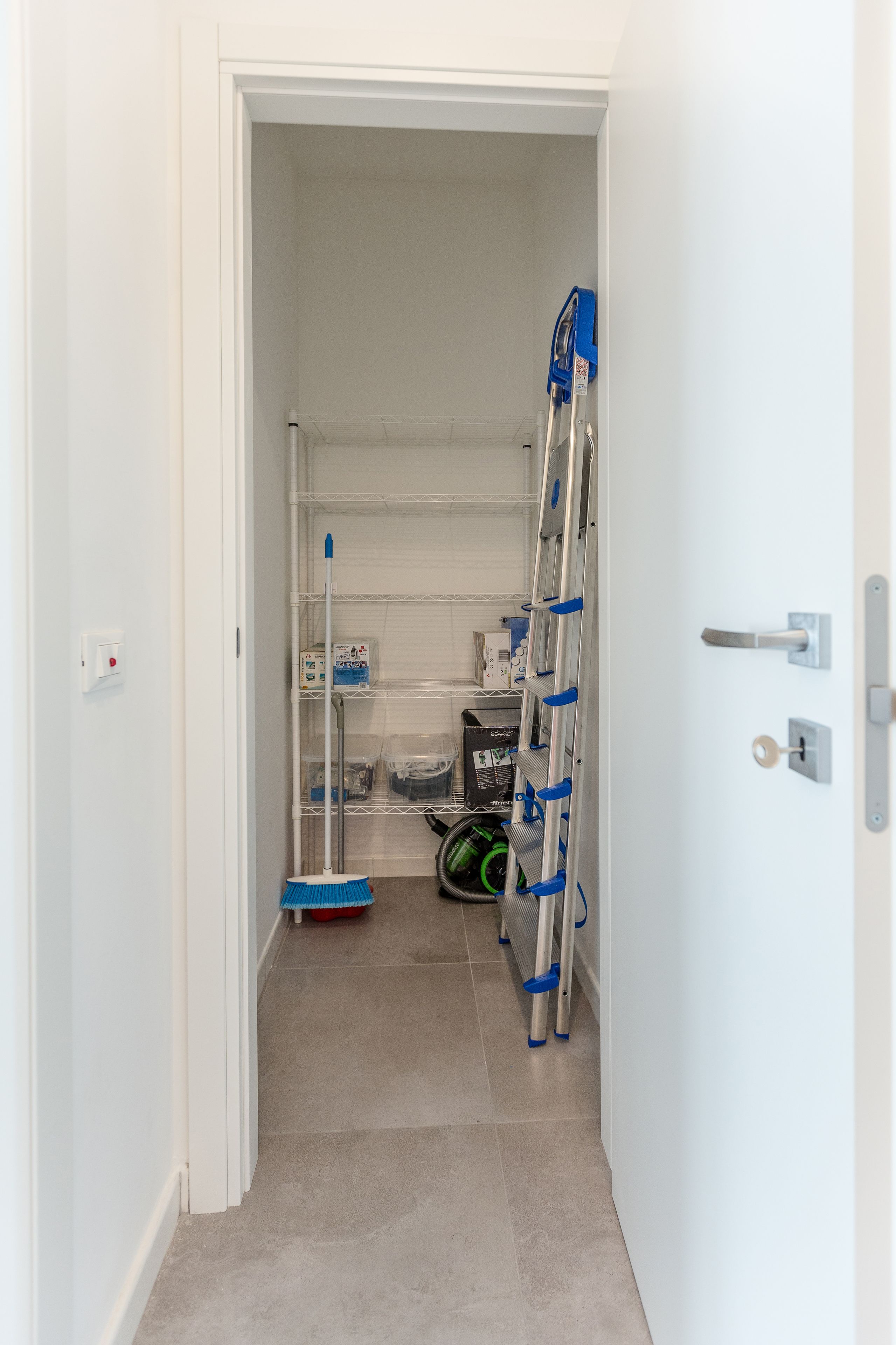 Een smalle gang met een witte deur, een metalen ladder en een bezem die tegen de muur staat.