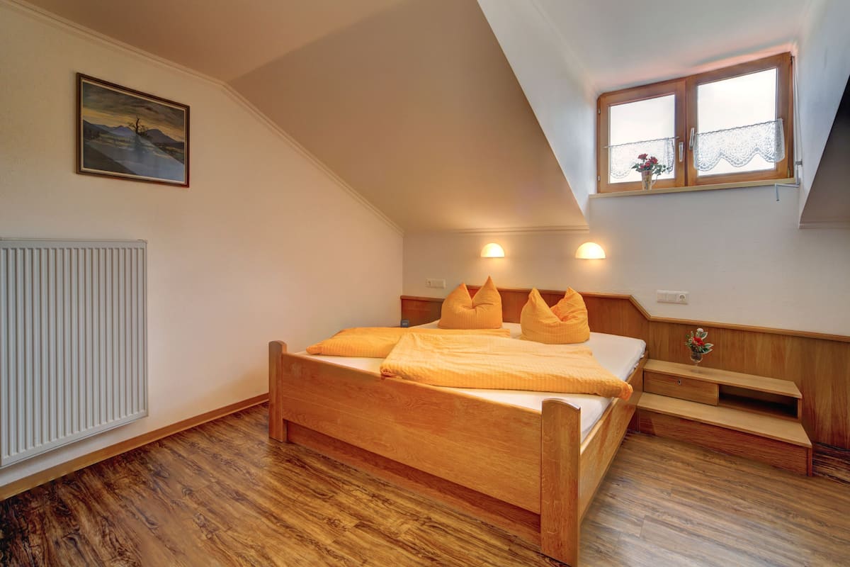 Ein Schlafzimmer mit einem Holzbett, einem Heizkörper, einem Gemälde, einem Fenster mit Spitzenvorhängen und einem Nachttisch mit einer Vase.