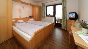 Quarto com cama de madeira, uma escrivaninha com cadeira, televisão e janela com cortinas.