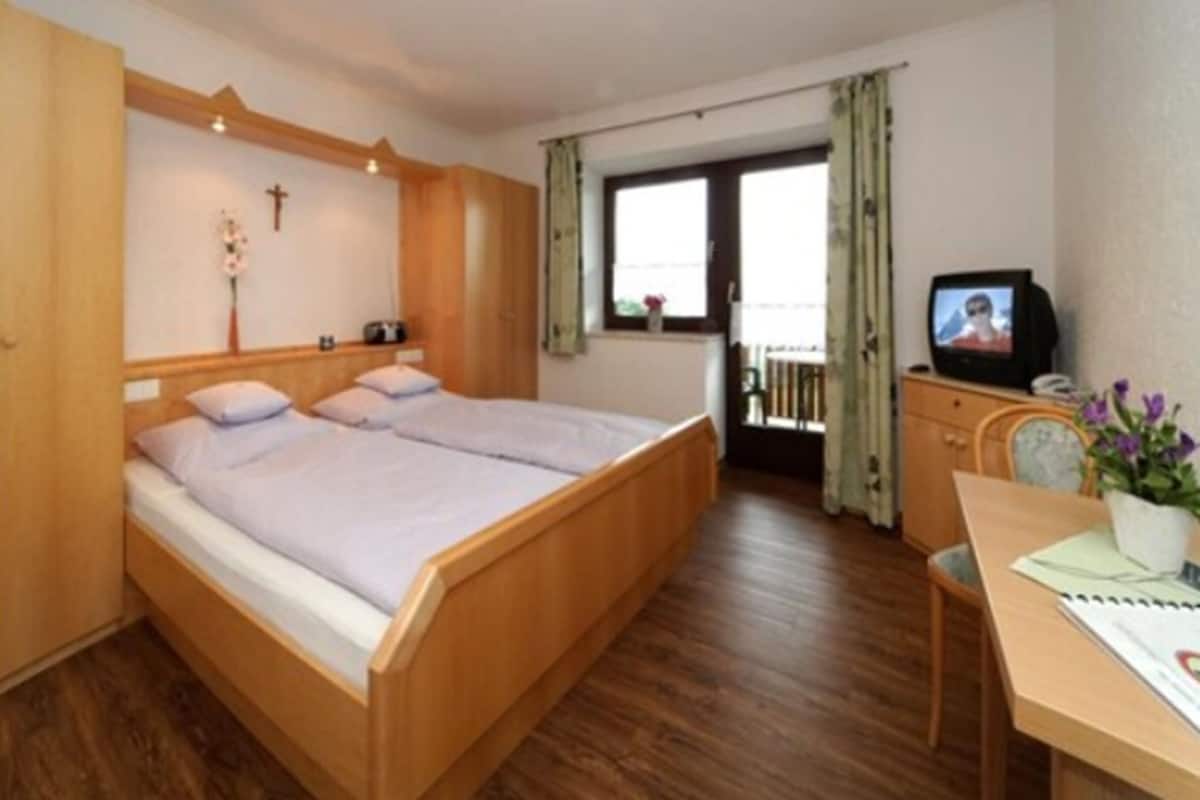 Ein Schlafzimmer mit einem hölzernen Bett, einem Schreibtisch mit Stuhl, einem Fernseher und einem Fenster mit Vorhängen.