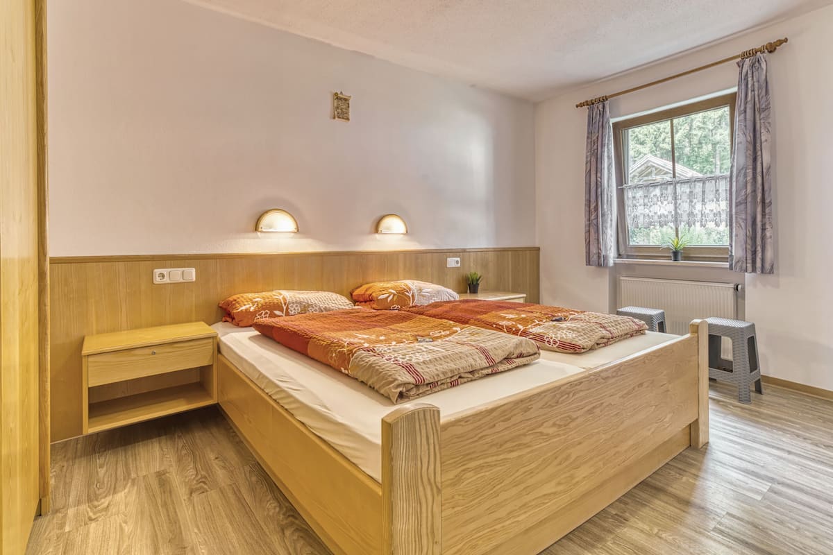 Ein Schlafzimmer mit einem hölzernen Bett, Nachttischen, einem Fenster mit Vorhängen und einem kleinen Hocker.