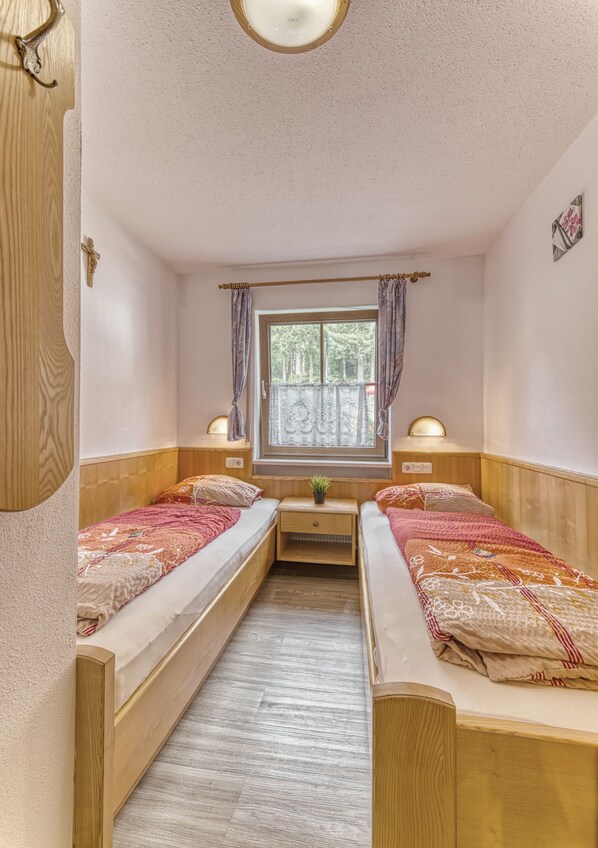 A small, cozy room with two wooden beds, a window with curtains, a small table with a lamp, and a wall-mounted shelf.