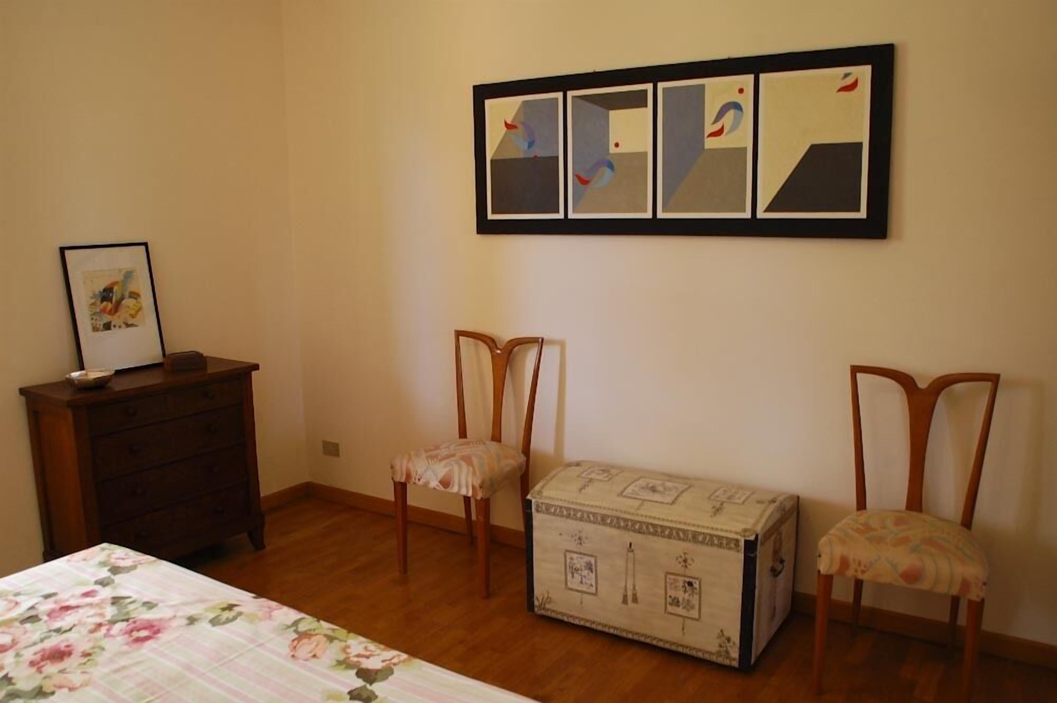 A room with a bed, a dresser, two chairs, a chest of drawers, and a framed abstract art piece on the wall.