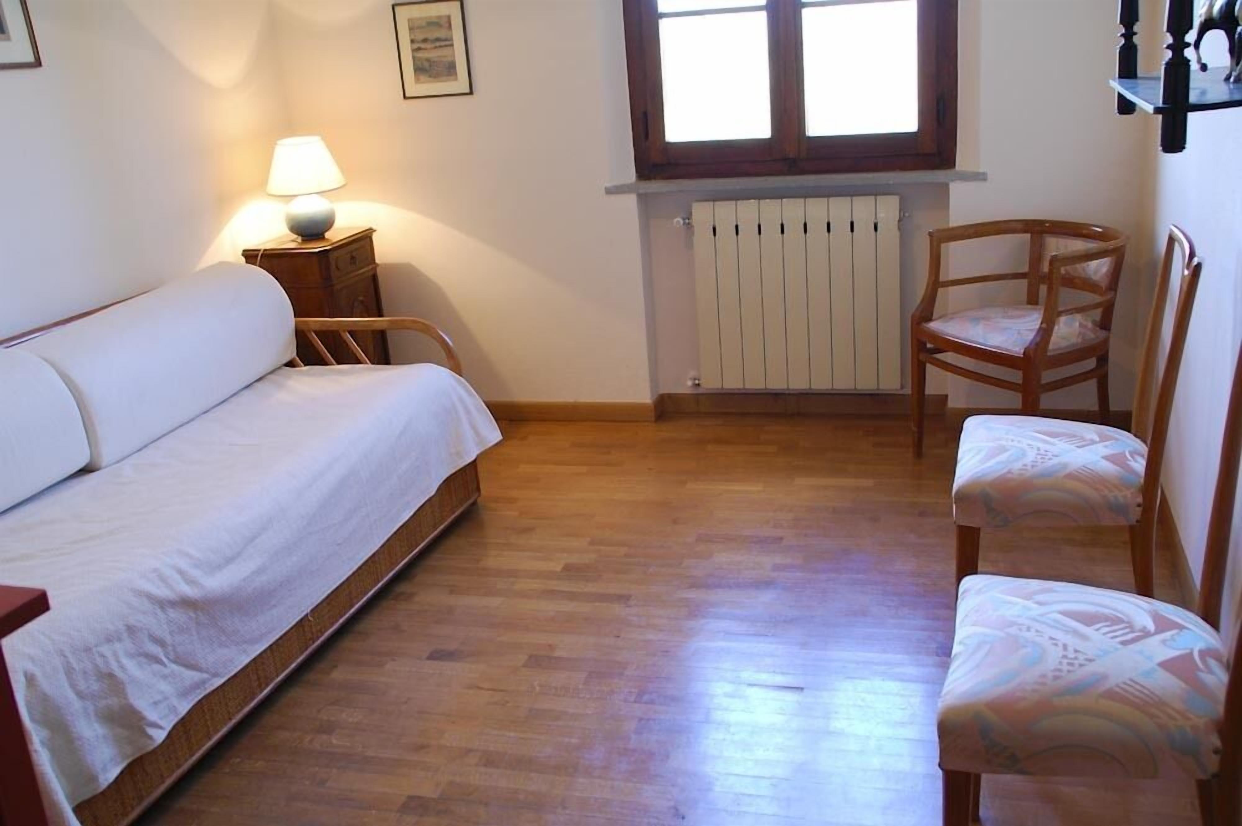 Un dormitorio con cama, una silla, una mesita de noche con lámpara, ventana y radiador.