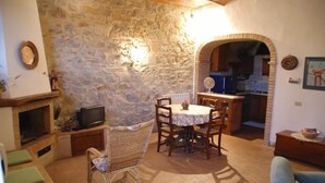 Un salon chaleureux avec un mur en pierre, une cheminée, une table à manger avec des chaises, et une cuisine visible à travers une arche.