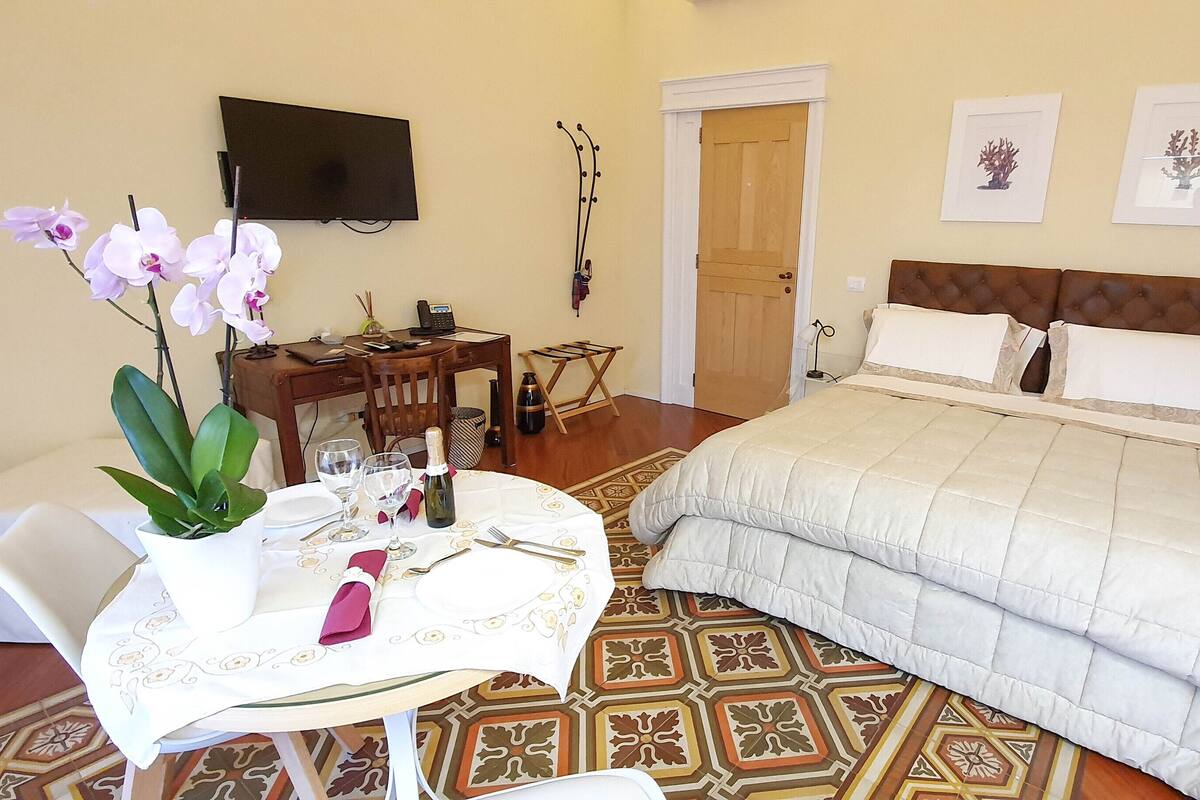 A hotel room with a bed, a desk with a chair, a small table with a plant, and a TV mounted on the wall.