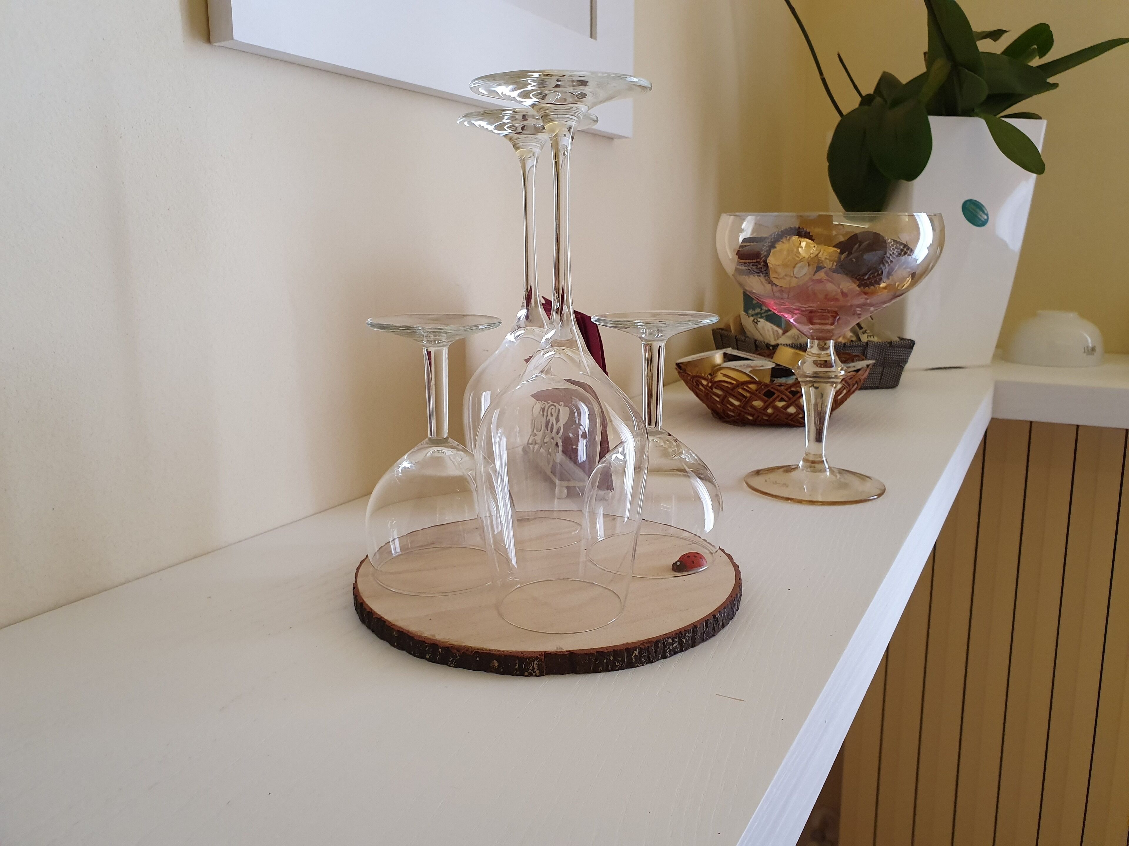 A clear glass wine rack with multiple glasses on a wooden base, a basket of snacks, and a potted plant on a white counter.