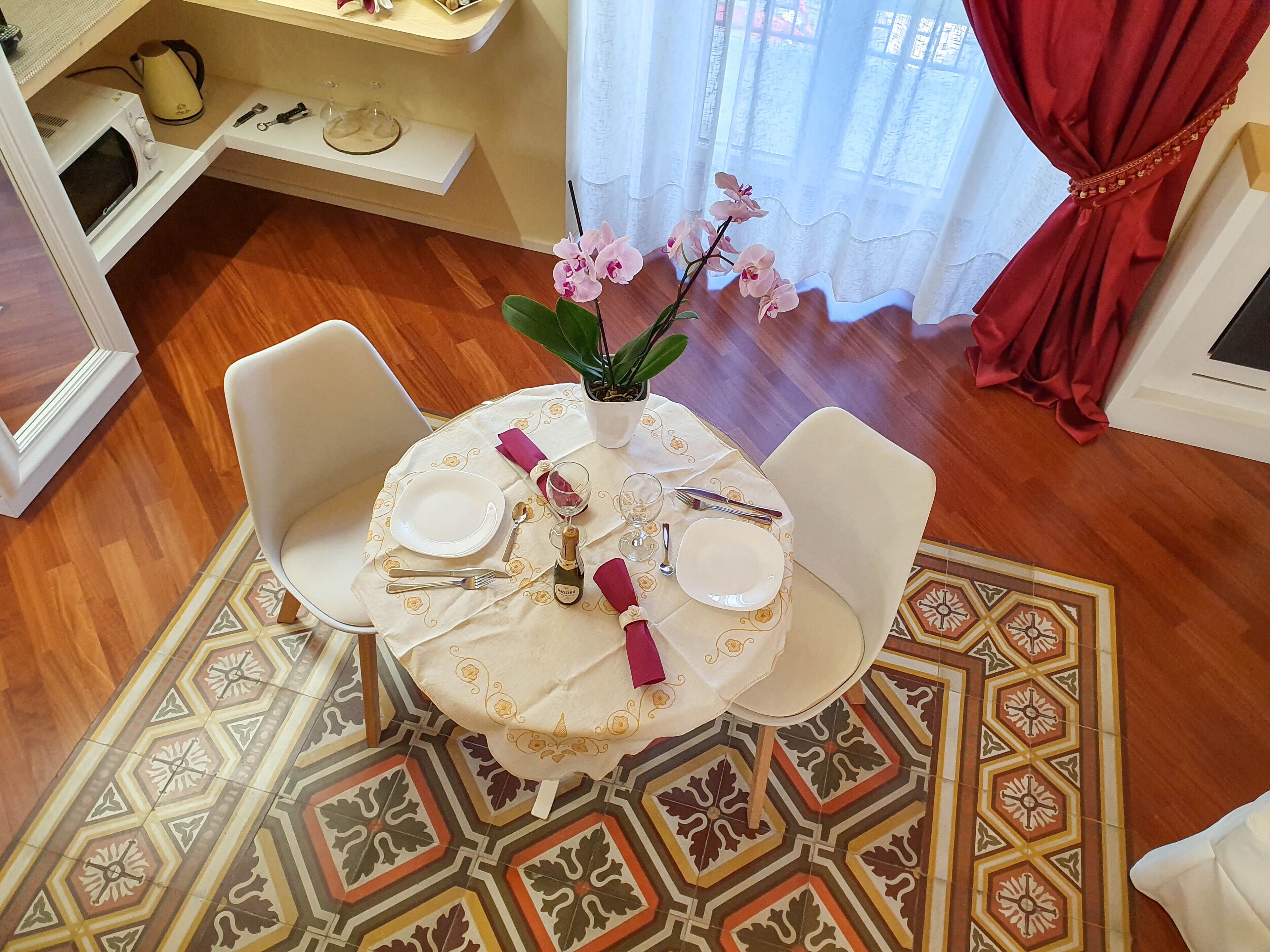 Un comedor con una mesa redonda dispuesta para dos personas, sillas blancas, un jarrón con flores rosadas y una alfombra estampada.