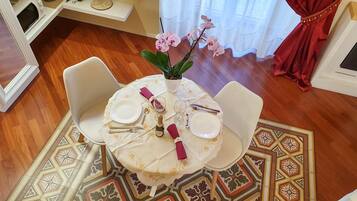 Un comedor con una mesa redonda dispuesta para dos personas, sillas blancas, un jarrón con flores rosadas y una alfombra estampada.