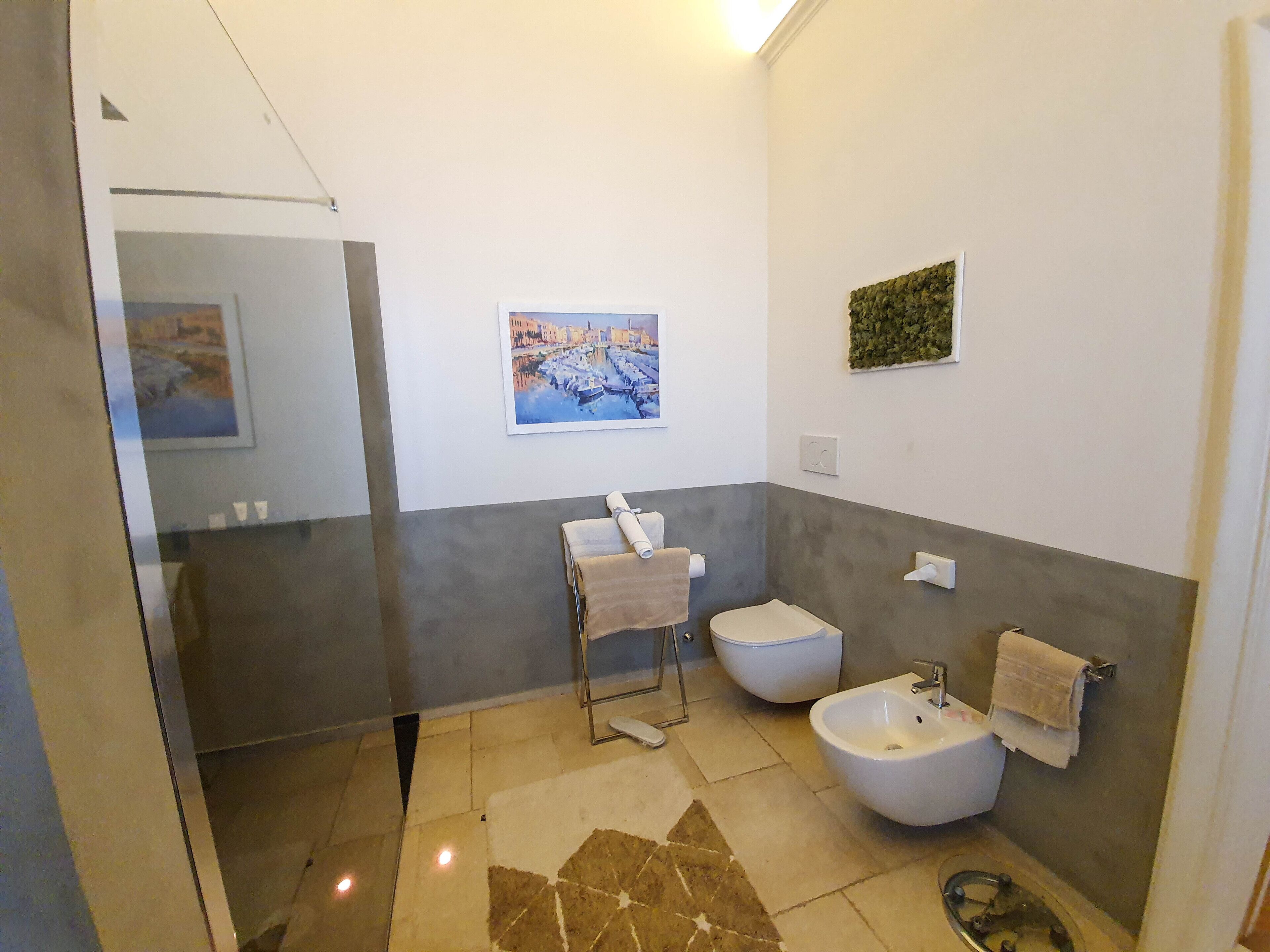 A modern bathroom with a shower, two bidets, a sink, and a toilet. There is a framed painting on the wall and a small green plant.