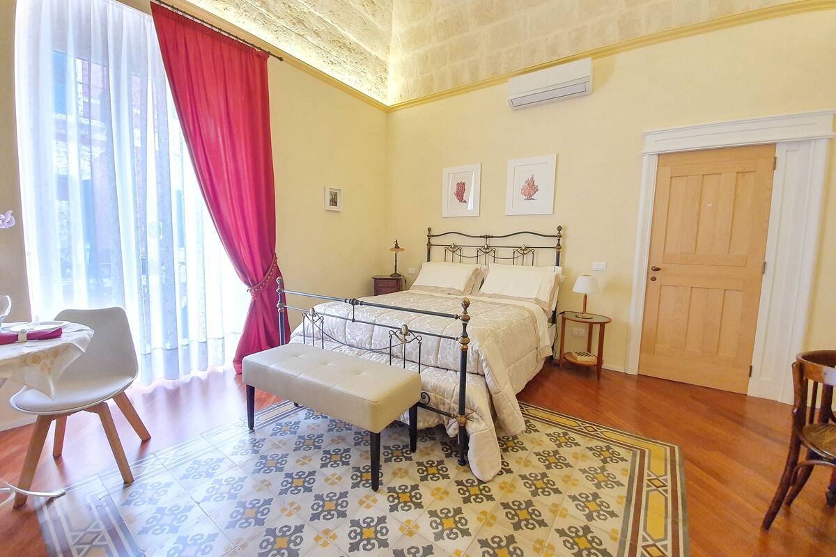 A bedroom with a bed, a bench, a chair, a bedside table, a lamp, and a door.