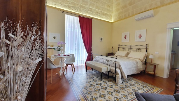 A bedroom with a bed, bedside table, lamp, and a small desk with a chair. There is a window with curtains and a view of the outside. - Turenum Apartment B&B (Trani)