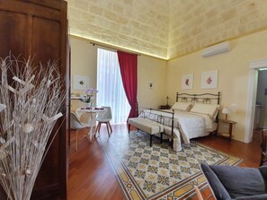 Quarto com cama, mesa de cabeceira, luminária e uma pequena escrivaninha com cadeira. Há uma janela com cortinas e vista para o exterior.