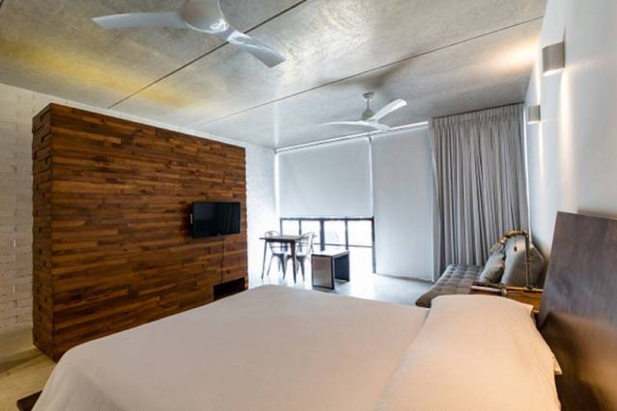 A modern hotel room with a large bed, a wooden accent wall, a flat-screen TV, a small table with chairs, and a large window with curtains.