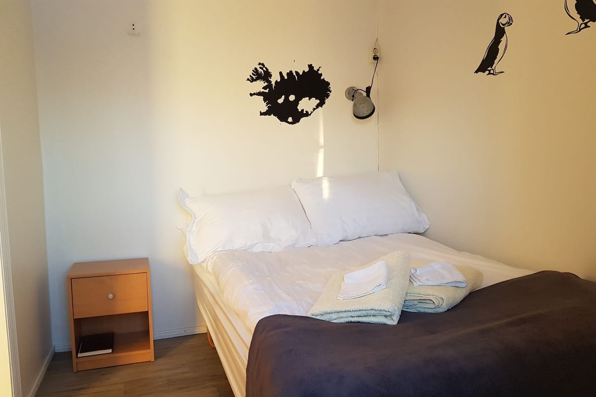 A neatly made bed with white linens and a folded towel, a wooden nightstand, and a wall-mounted light fixture.