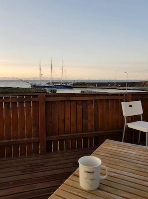 A wooden deck with a table and a chair, overlooking a body of water with a sailboat and mountains in the distance.