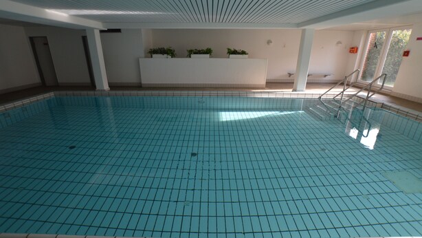 Una piscina interna con pavimento piastrellato, una reception in bianco con piante e una finestra che si affaccia su alberi.