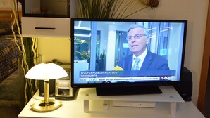Un televisore che mostra un notiziario, un mobile porta televisione bianco con una lampada e oggetti decorativi sugli scaffali.