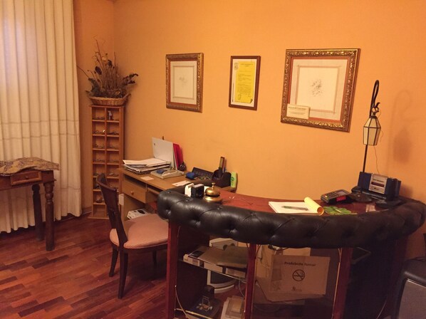 Una habitación con escritorio de madera, una silla, una laptop, una lámpara y diplomas enmarcados en la pared.