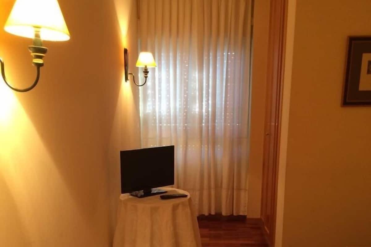 Ett hotellrum med trägolv, ett litet runt bord med en lampa, en säng med randiga sängkläder och ett fönster med tunna, genomskinliga gardiner.