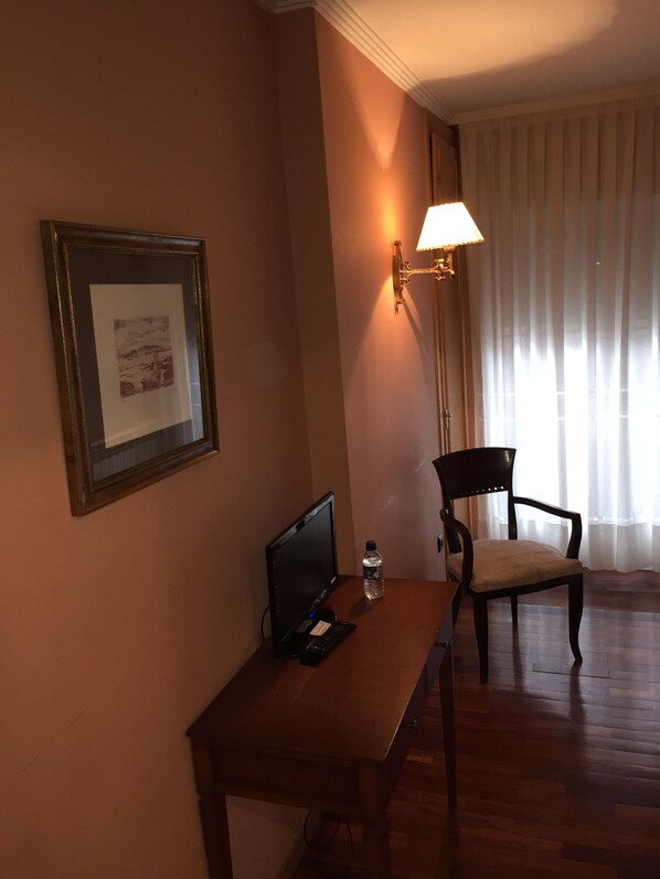 A hotel room with a wooden desk, a laptop, a framed picture, a chair, a lamp, and a window with curtains.