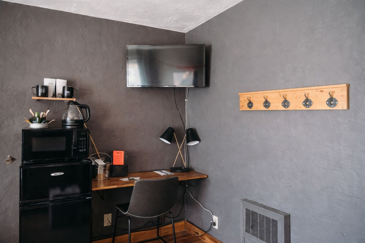 A compact kitchenette with a microwave, kettle, and coffee maker. A flat-screen TV is mounted above a wooden shelf with hooks.