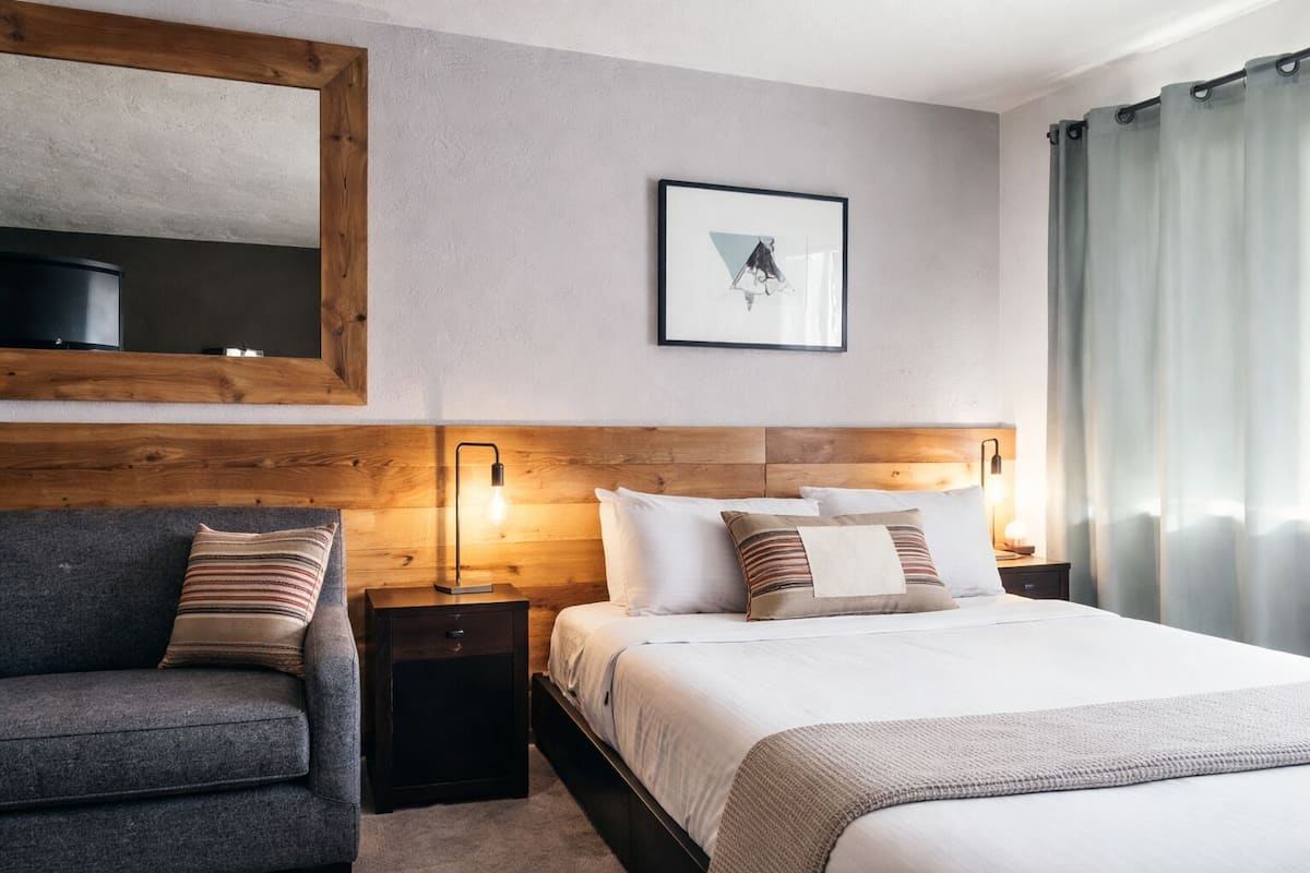 A hotel room with a bed, a nightstand, a lamp, a chair, a mirror, and a picture on the wall.