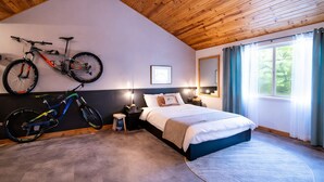 A bedroom with a bed, two bicycles, a nightstand, a lamp, a window with curtains, and a wooden ceiling.