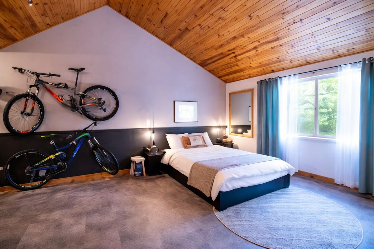 A bedroom with a bed, two bicycles, a nightstand, a lamp, a window with curtains, and a wooden ceiling.