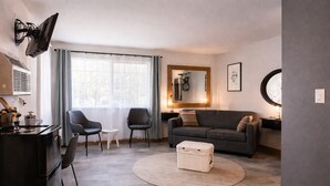 A modern living room with a sofa, two chairs, a small table, a TV mounted on the wall, and a large window with curtains.